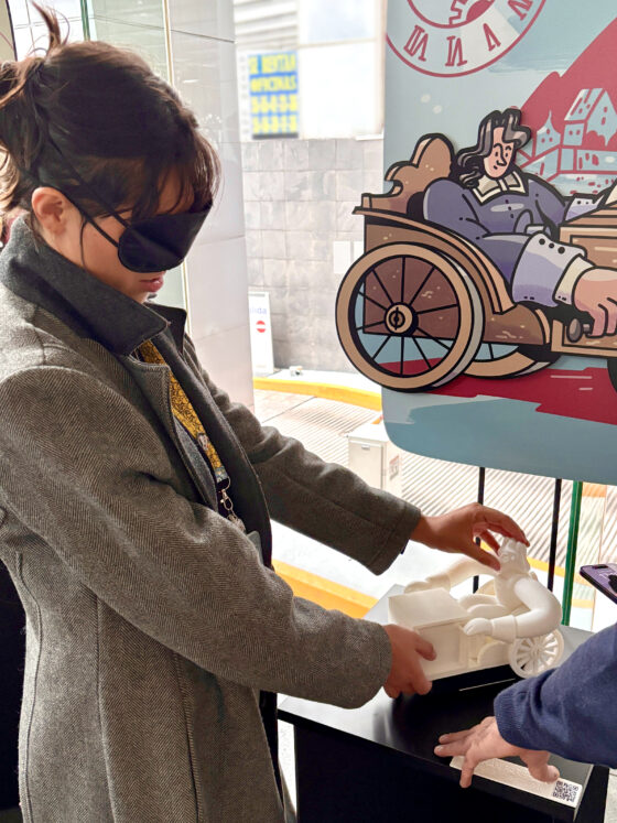 Foto vertical en perspectiva de una mujer con los ojos vendados tocando la réplica táctil del relojero Stephan Farffler. La réplica está montada sobre un pedestal negro, y al fondo se ve una ilustración de gran formato del mismo personaje.