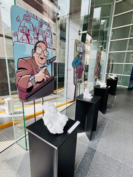 Foto vertical en perspectiva de la exposición "los muros que derribamos" instalada en el lobby de un corporativo. Se observan 5 módulos rectangulares de color negro, sobre los que se exhiben dibujos de personajes y sus respectivas réplicas táctiles impresas en 3D.