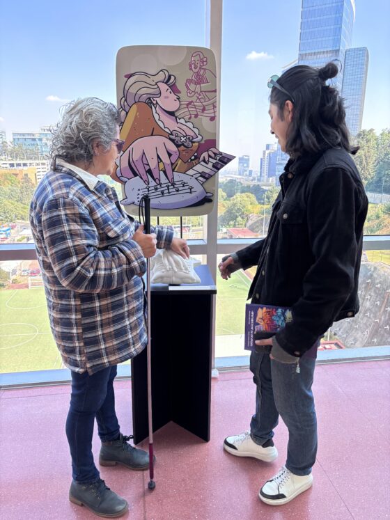 Foto vertical de una mujer con bastón blanco frente a un pedestal que tiene una ilustración de Maria Theresia Von Paradis, una pianista con discapacidad visual, y su respectiva réplica impresa en 3D. Frente a la mujer hay un guía con cabello largo explicándole la pieza.